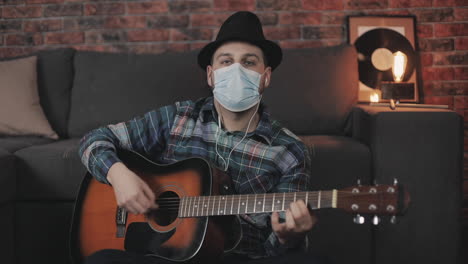 Portrait-Of-Young-Male-Musician-With-Medical-Face-Mask-Playing-Guitar,-Looking-At-Camera,-At-Home-During-Lockdown-Due-To-The-Covid-19-Pandemic