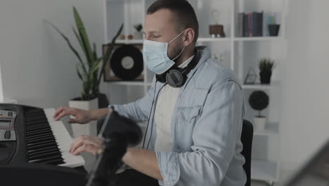 Male-Musician-With-Medical-Face-Mask-Playing-Electric-Keyboard-And-Using-A-Laptop-At-Home-During-Lockdown-Due-To-The-Covid-19-Pandemic