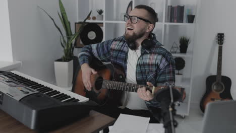 Male-Musician-Singing-And-Playing-Guitar-Next-To-His-Electric-Keyboard-At-Home