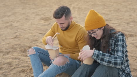 Young-female-and-male-friends-eating-take-away-food-sitting-on-the-beach.