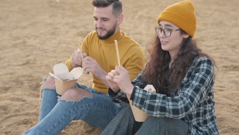 Young-Female-And-Male-Friends-Eating-Take-Away-Food-Sitting-On-The-Beach