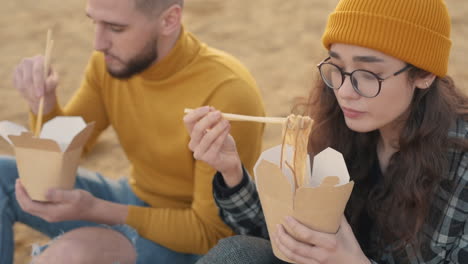 Young-Female-And-Male-Friends-Eating-Take-Away-Food-Sitting-On-The-Beach-1