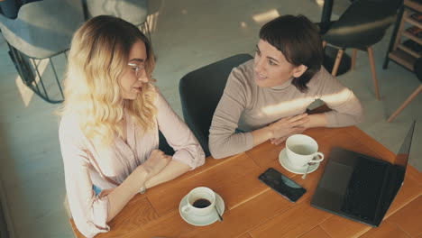Blonde-And-Brunette-Female-Friends-With-A-Laptop-And-A-Smartphone-Talking-In-A-Coffee-Shop
