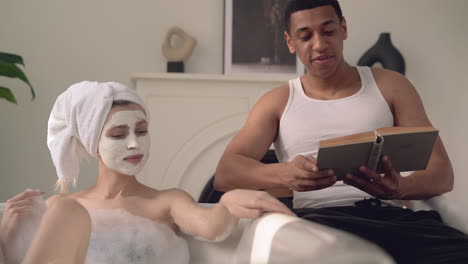 Woman-With-Face-Mask-Taking-A-Relaxing-Bath,-Black-Man-Reads-A-Book