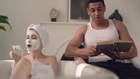 Female-With-Face-Mask-Taking-A-Relaxing-Bath,-Black-Male-Reads-A-Book