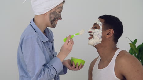 Female-With-Facial-Mask-Applying-Scrub-To-A-Black-Male