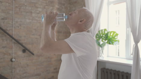Old-Male-Drinks-Water-And-Rests-After-Exercising-And-Stretching-At-Home