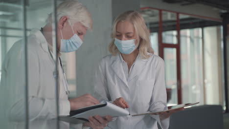 A-Pair-Of-Doctors-With-Masks-Compare-Results-From-A-Notebook-And-Tablet