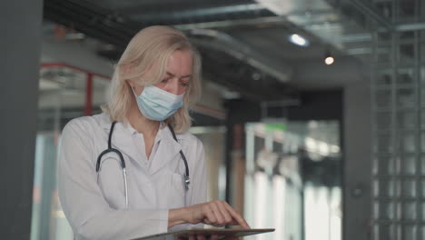 A-Blonde,-Middle-Aged-Female-Doctor-In-A-Facemask-Consults-Data-On-A-Tablet