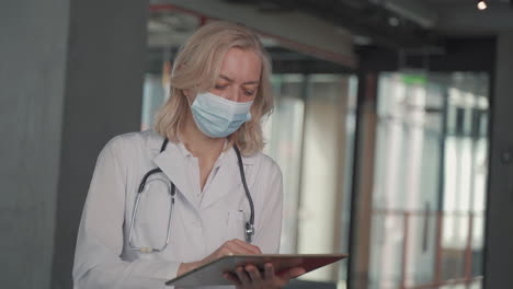 A-Blonde,-Middle-Aged-Female-Doctor-In-A-Facemask-Consults-Data-On-A-Tablet-1