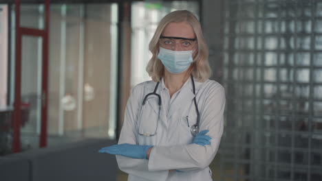 A-Blonde,-Middle-Aged-Female-Doctor-In-A-Facemask-Looks-Directly-At-The-Camera-With-Crossed-Arms-1