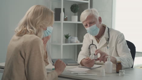 A-Grey-Haired,-Middle-Aged-Doctor-In-A-Facemask-Prescribes-Medicines-To-A-Female-Patient