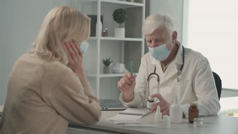 A-Grey-Haired,-Middle-Aged-Doctor-In-A-Facemask-Prescribes-Medicines-To-A-Female-Patient-2