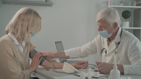 A-Grey-Haired,-Middle-Aged-Doctor-In-A-Facemask-Doctor-Takes-Blood-Pressure-To-A-Female-Patient