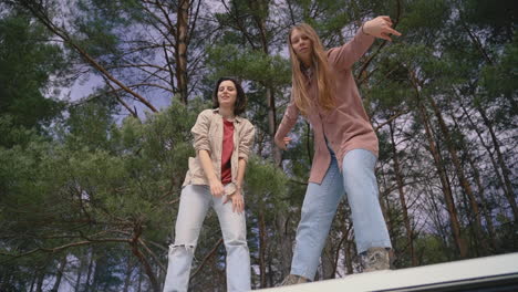 Two-Young-Girl-Friends-Make-Fools-Of-Themselves-On-Top-Of-The-Roof-Of-A-Caravan-In-The-Middle-Of-The-Forest-Looking-At-The-Camera