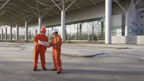 Construction-Workers-In-Orange-Uniform-And-Hardhats-Looking-Over-Plans-Together-1