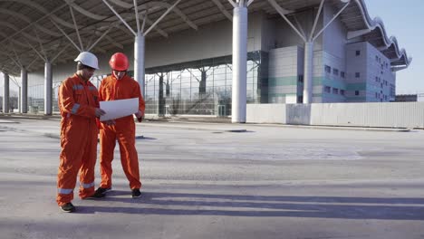 Two-Construction-Workers-In-Orange-Uniform-And-Helmets-Meeting-Each-Other-At-The-Bulding-Object,-Shaking-Hands-And-Examining-The-Constructed-Building-Together