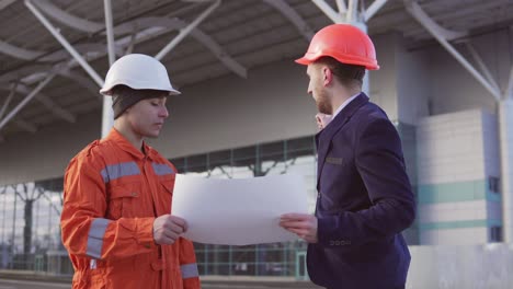 Young-Manager-Of-The-Project-In-A-Black-Suit-Examining-The-Building-Object-With-Construction-Worker-In-Orange-Uniform-And-Helmet