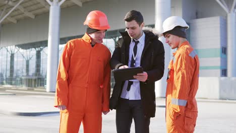 Investor-Des-Projekts-In-Einem-Schwarzen-Anzug,-Der-Das-Bauobjekt-Mit-Bauarbeitern-In-Orangefarbener-Uniform-Und-Helmen-Untersucht