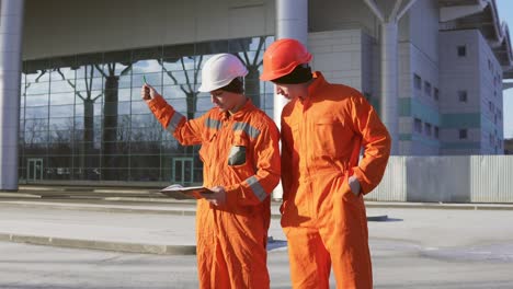 Zwei-Bauarbeiter-In-Oranger-Uniform-Und-Helmen-Schauen-Sich-Gemeinsam-Pläne-An