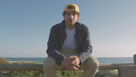 Handsome-Man-In-Cap-Sitting-On-Boardwalk-Looking-At-Camera