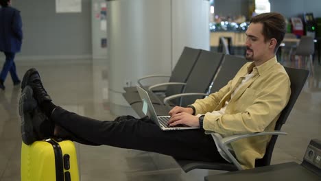 Un-Joven-Aburrido-Está-Sentado-En-El-Aeropuerto,-Esperando-Que-Salga-El-Avión-Y-Usando-Una-Computadora-Portátil