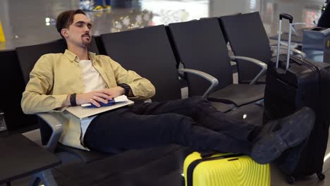 A-Bearded-Man-In-Yellow-Shirt-Sleeping-Holding-Legs-On-The-Yellow-Suitcase,-Holding-His-Stuff-In-The-Airport-Lounge-While-Sitting