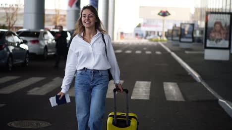 Excited,-Stylish-Woman-Walk-With-Trolley-Yellow-Case-By-Empty-Airport-Terminal-Outside-Road,-Slender-Female-Wearing-Jeans-And-White-Shirt