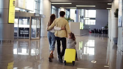 Back-View-Of-Modern-Family-Mother,-Father,-Daughter-Walking-The-Airport-Hall-And-Riding-Their-Daughter-On-Suitcase-Before-The-Departure-To-The-Vacation