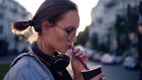 Side-View-Of-A-Beautiful-Young-Woman-With-Headphones-On-Her-Neck-Drinking-Beverage-Using-A-Straw