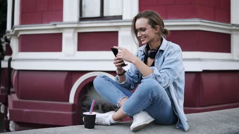 A-Fashionable-Girl-In-Glasses-Sits-On-The-Street-On-A-Parapet,-Concentrates-On-Her-Mobile-Phone