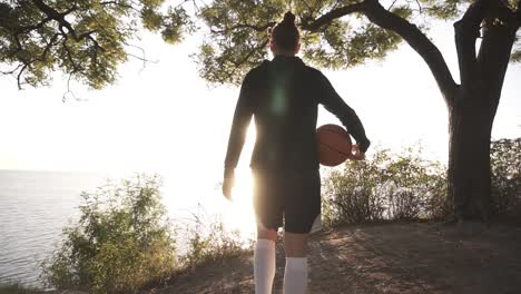 A-Basketball-Player-Girl-Comes-With-A-Ball-In-Her-Hand,-Coming-Up-To-The-Slope-With-Trees-Around