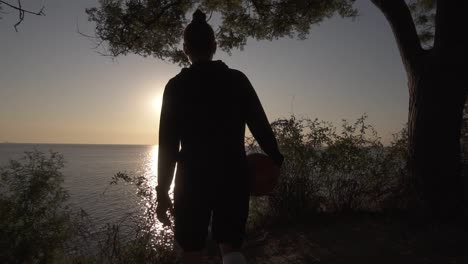 A-Silhouette-Of-A-Basketball-Player-Girl-Comes-With-A-Ball-In-Her-Hand,-Coming-Up-To-The-Slope-With-Trees-Around
