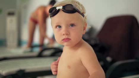 Portrait-Of-A-Beautiful-Toddler-With-Goggles-Sitting-By-The-Pool-And-Looking-In-The-Camera-Waiting-For-His-Mother-To-Come