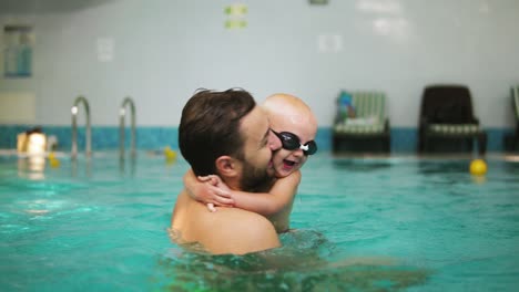 Young-Father-Lifting-His-Little-Boy-In-Protective-Glasses-From-The-Water-While-Teaching-Him-How-To-Swim-In-The-Swimming-Pool