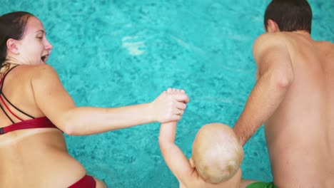 Back-View-Of-Young-Mother-And-Father-Together-With-Their-Child-Jumping-In-The-Swimming-Pool-Holding-Hands