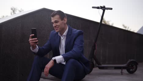 Attractive-Businessman-Sitting-On-The-Stairs-And-Does-Video-Cahtting-On-Smartphone-Outside