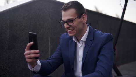 Excited,-Smiling-Businessman-Sitting-On-The-Stairs-And-Does-Video-Cahtting-On-Smartphone-Outside