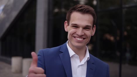 Upwards-Footage-Of-A-Young-Businessman-In-Elegant-Blue-Suit-Standing-Outdoors,-Cheerfully-Smiling-And-Showing-Thumb-Up