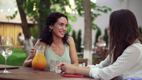 People,-Communication-And-Friendship-Concept-Smiling-Young-Women-Drinking-Orange-Juice-And-Talking-At-Outdoor-Cafe-On-Summer