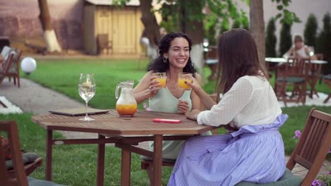 People,-Communication-And-Friendship-Concept-Smiling-Young-European-Women-Drinking-Orange-Juice-At-Outdoor-Cafe,-Clinking-Glasses