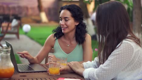 Two-Women-Sitting-At-The-Outdoors-Cafe-Talking,-Sharing-News,-Brunette-Girl-Showing-Something-On-Laptop-Screen-And-Laughing