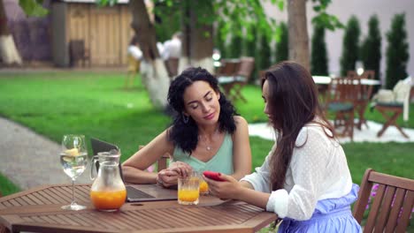 Two-Women-Sitting-At-The-Outdoors-Cafe-Talking,-Sharing-News,-Girls-Showing-Something-On-Laptop-Screen-And-Smartphone