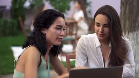 Two-Women-Sitting-At-The-Outdoors-Cafe-Talking,-Sharing-News,-Brunette-Girl-Showing-Something-On-Laptop-Screen,-Gesturing