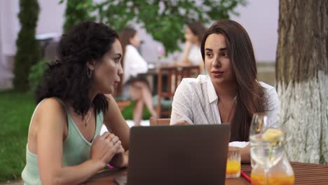 Two-Women-Sitting-At-The-Outdoors-Cafe-Talking,-Sharing-Ideas,-Showing-Something-On-Laptop-Screen,-Gesturing,-Arguing