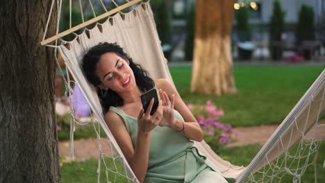 Portraitof-A-Woman-Relaxing-In-A-Hammock-Outdoors,-Surfing-Internet-In-Her-Smartphone,-Clicks-On-The-Screen-And-Smiling