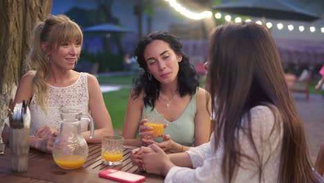 Three-Elegant-Female-Friends-Sit-In-The-Outdoors-Cafe,-Drink-Juice-And-Have-Fun-Communicating
