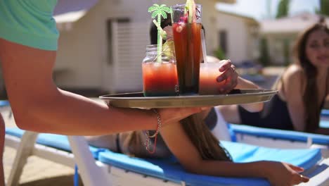 An-Unrecognizable-Waiter-Bringing-Cocktails-For-Beautiful-Young-Girls-Relaxing-By-The-Pool