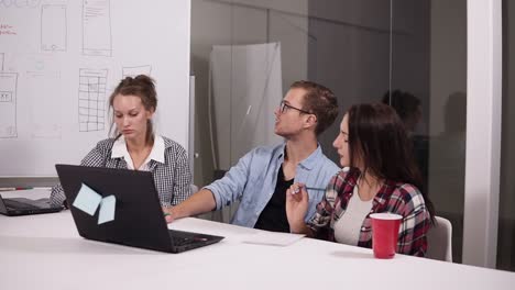 Young-Man-In-Glasses-And-Two-Women-Discussing-Something-With-Each-Other-While-Sitting-At-The-Office-Table-In-Creative-Workplace