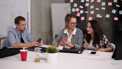 Group-Of-Three-Young-Coworkers-Sitting-In-The-Modern-Loft-Office-White-Huge-Table,-Looking-At-Their-Collegue-Smartphone,-Discussing-Something